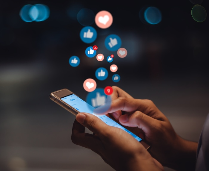 Close-up of a person holding a smartphone with floating social media icons, including likes, hearts and notifications, above the screen.